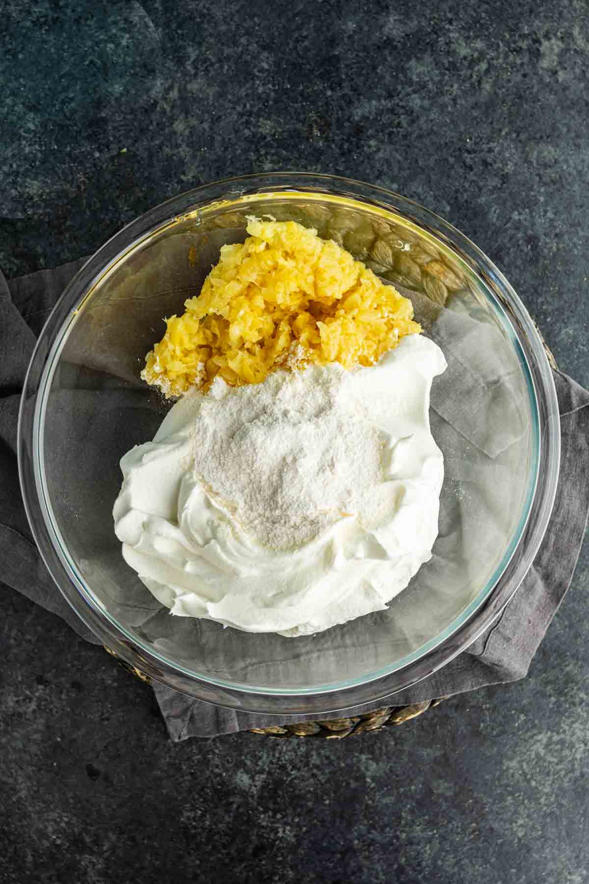A glass bowl containing crushed pineapple, whipped topping, and powdered pudding mix—a classic combination for a Southern dessert like the famous Pig Pickin’ Cake—sits on a gray napkin over a dark surface.