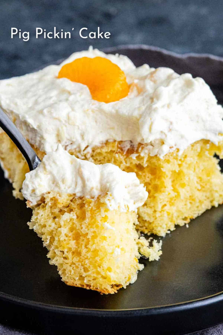A slice of yellow cake with whipped cream topping and a mandarin orange segment on a black plate, with a fork taking a piece. Text on image reads “Pig Pickin’ Cake.”.
