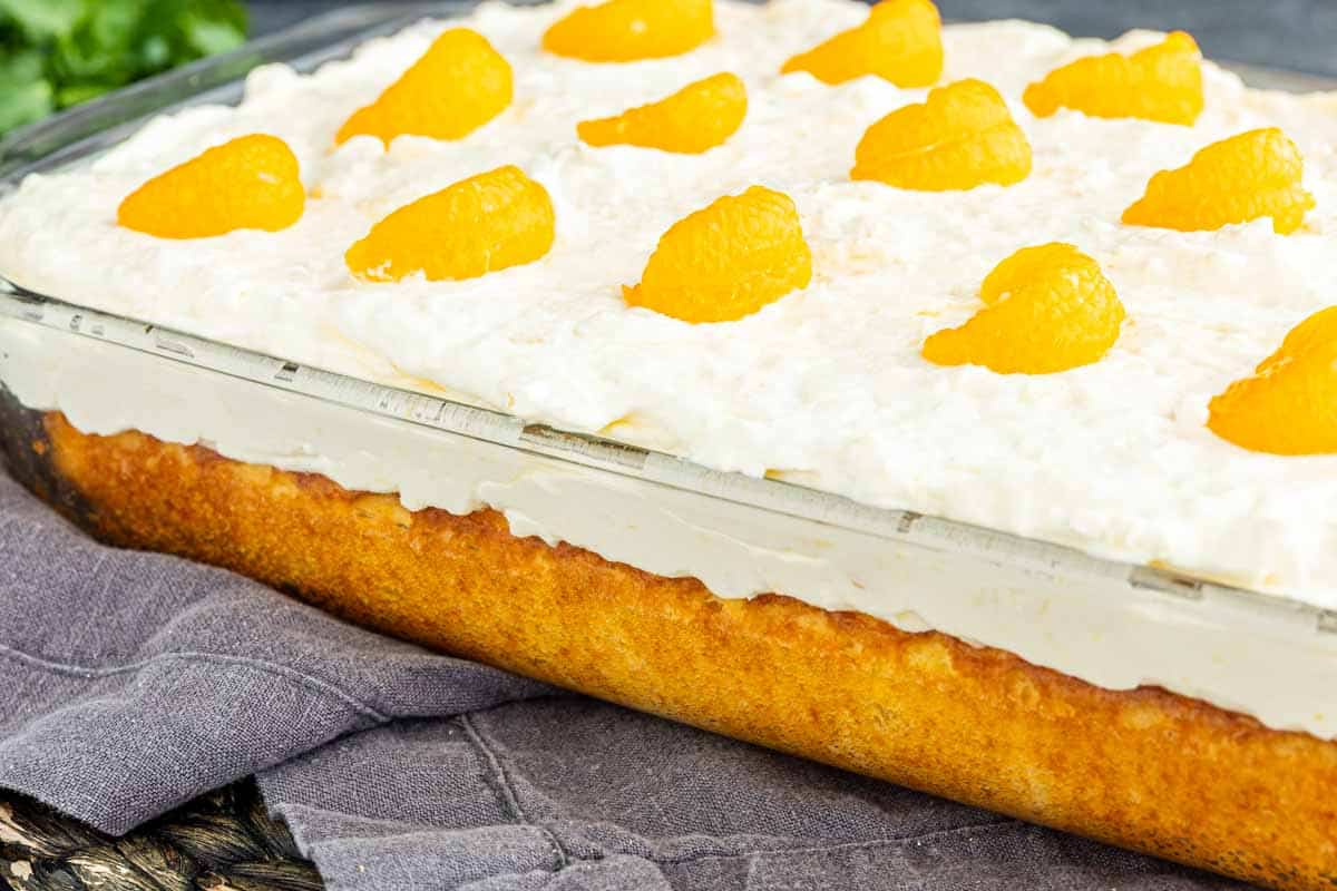 A rectangular glass dish filled with a layered Southern dessert, topped with whipped cream and mandarin orange segments, sits on a light-colored cloth—reminiscent of a classic mandarin orange cake.