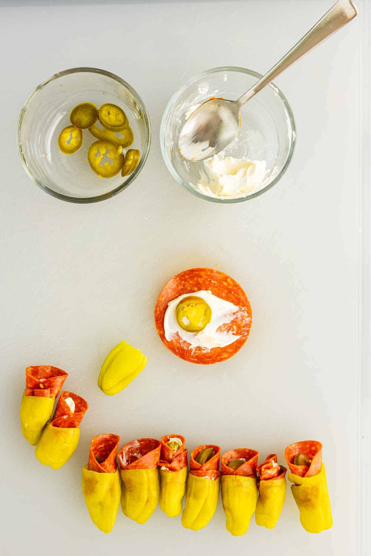 Two glass bowls with sliced jalapeños and cream cheese, a slice of pepperoni with cream cheese and a jalapeño on top, and several assembled pepperoni stuffed pepperoncinis snacks on a white surface.