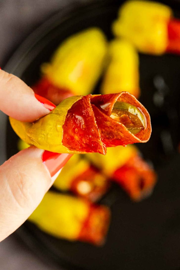 A hand holding a yellow pepper stuffed with cheese and wrapped in pepperoni, with more stuffed peppers on a plate in the background.
