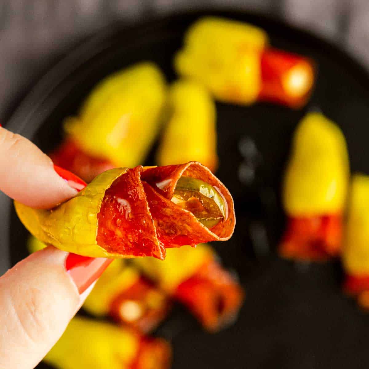 A hand holds a yellow pepperoni stuffed pepperoncini filled with sliced meat, with more air fryer stuffed pepperoncinis arranged on a black plate in the background.