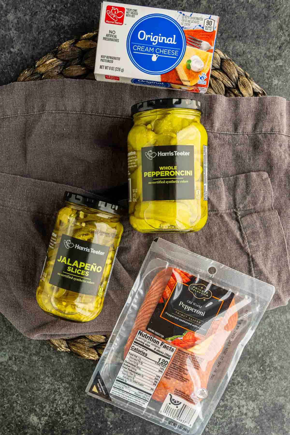 A package of cream cheese, two jars of pickled peppers, and a package of sliced pepperoni arranged on a gray cloth and woven mat—perfect ingredients for air fryer stuffed pepperoncinis.