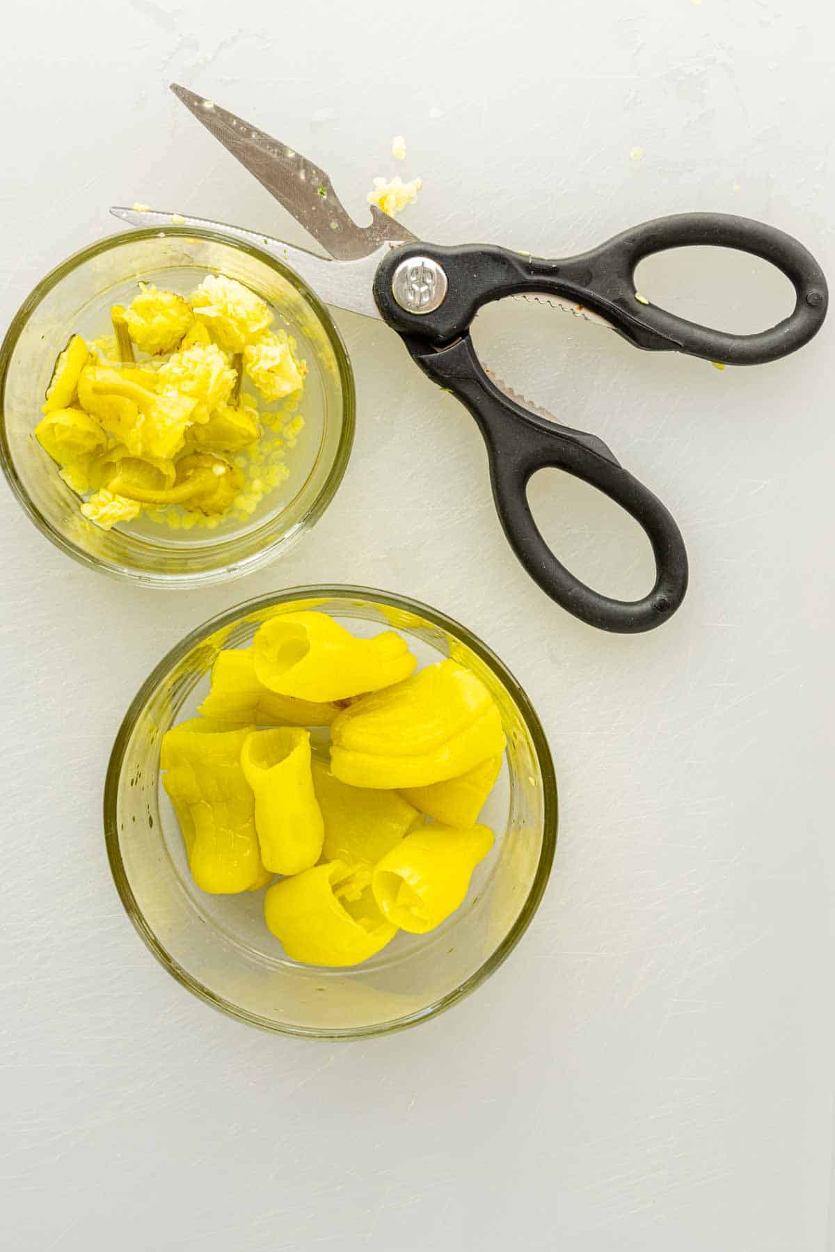 Two glass bowls with chopped yellow pickled peppers, ideal for making pepperoncini poppers or air fryer stuffed pepperoncinis, sit beside a pair of black kitchen scissors on a white surface.