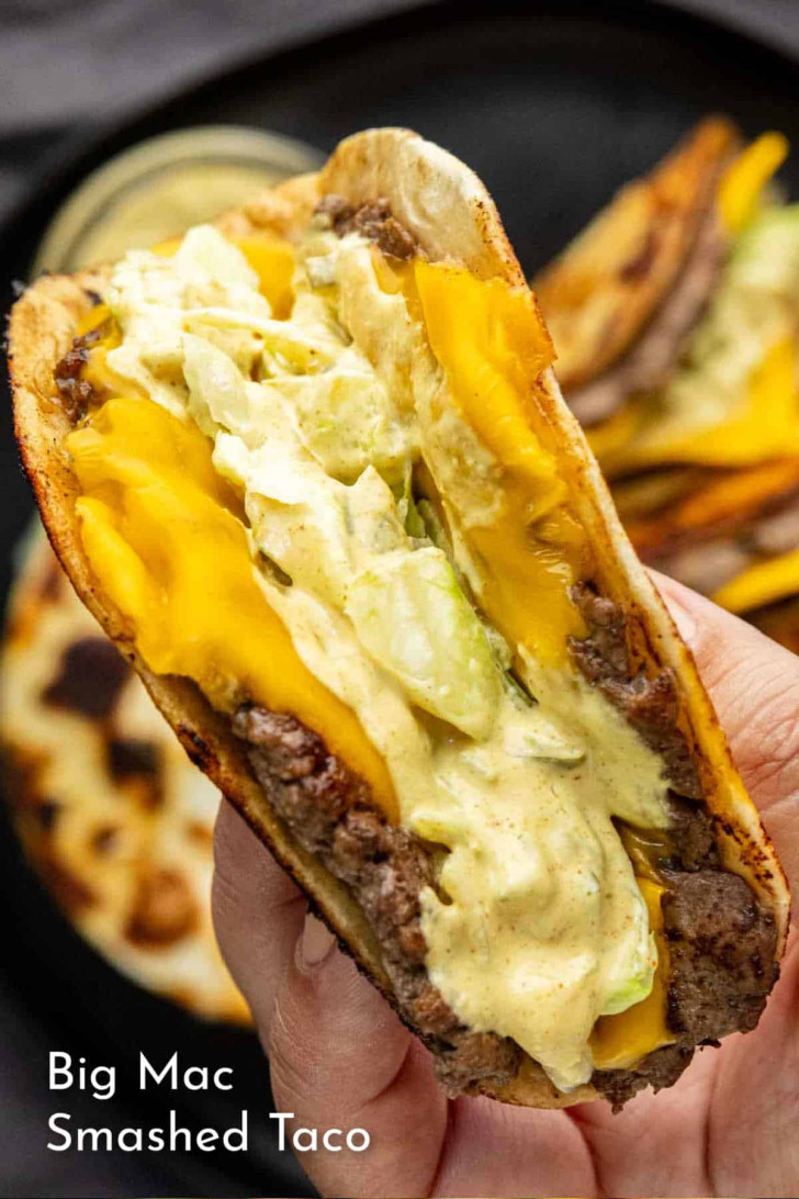 A hand holding a taco with ground beef, melted cheddar cheese, lettuce, and creamy sauce in a folded tortilla.
