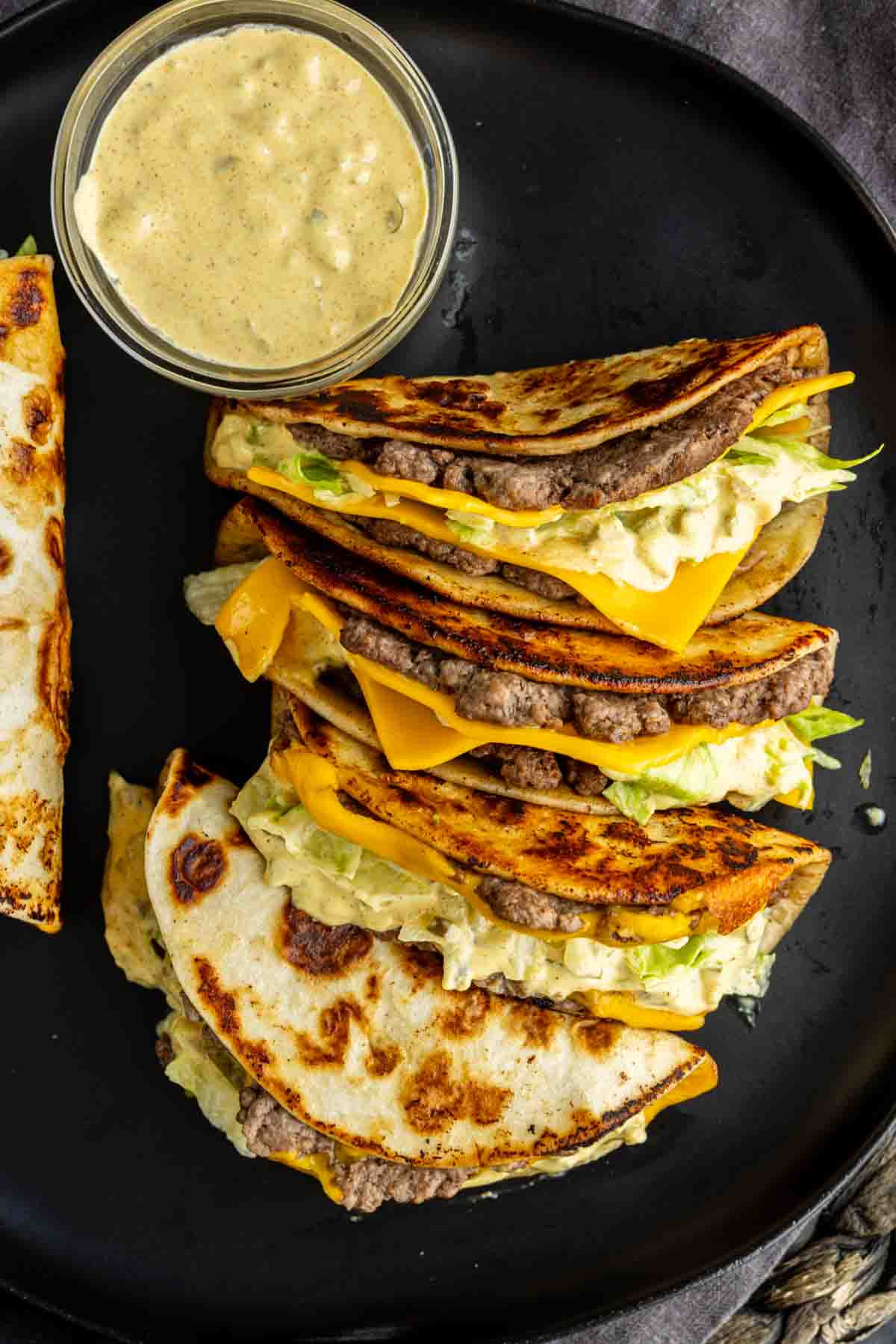 Three folded tortillas filled with ground beef, cheddar cheese, lettuce, and sauce are arranged on a black plate next to a small bowl of creamy dipping sauce—an easy weeknight dinner inspired by the viral taco recipe trend.