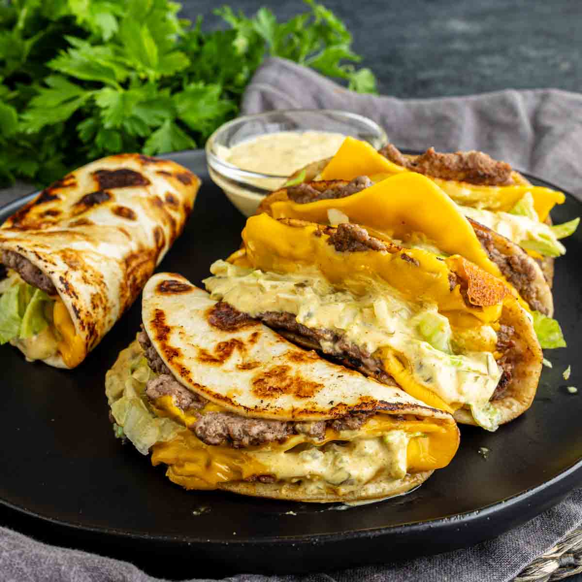 Three cheeseburger tacos filled with ground beef, cheddar cheese, lettuce, and sauce are on a black plate next to a small bowl of dip and fresh parsley—this viral taco recipe is perfect for an easy weeknight dinner.
