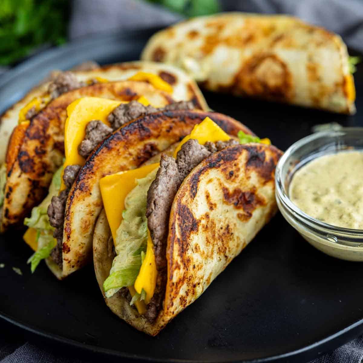 Three grilled tacos filled with beef, lettuce, and cheddar cheese are arranged on a black plate next to a small bowl of dipping sauce—an easy weeknight dinner inspired by the viral taco recipe trend.