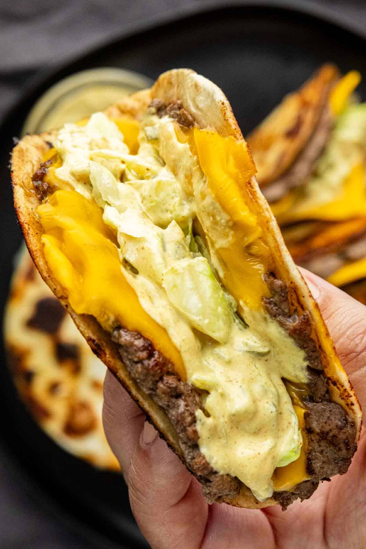 A hand holds a folded flatbread filled with beef, cheddar cheese, lettuce, and a creamy sauce—an irresistible take on the viral taco recipe—with more sandwiches on a black plate in the background.