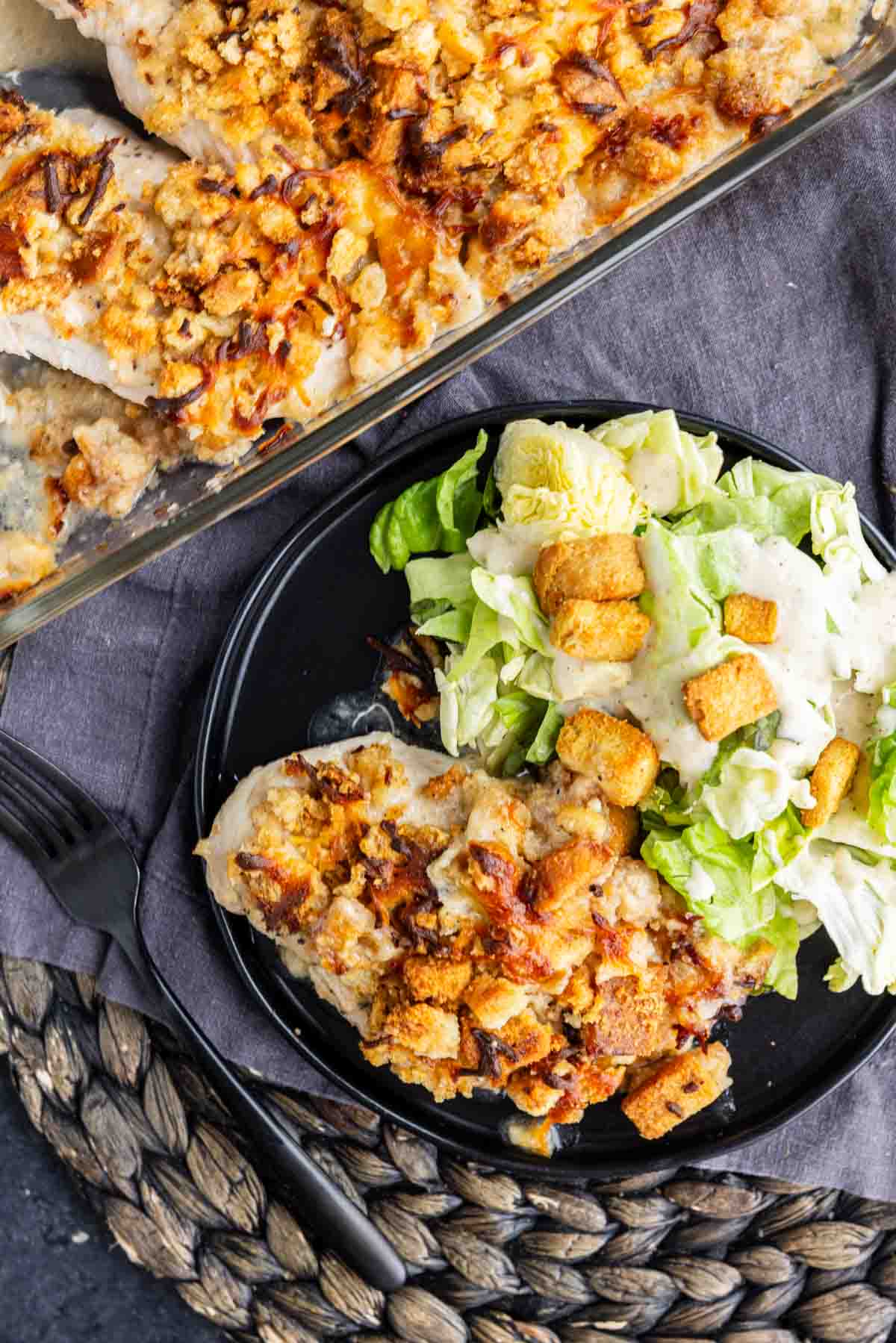 A plate with a serving of baked Caesar chicken topped with crumbs and melted cheese, alongside a green salad with croutons and dressing. A casserole dish with more easy weeknight chicken recipe is nearby.