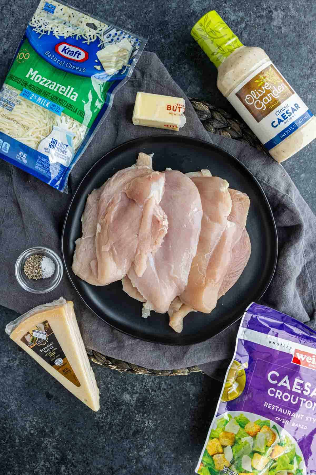 A plate of raw chicken breasts, perfect for baked Caesar chicken or any easy weeknight chicken recipe, is surrounded by mozzarella cheese, Caesar dressing, croutons, Parmesan cheese, butter, and black pepper on a dark surface.