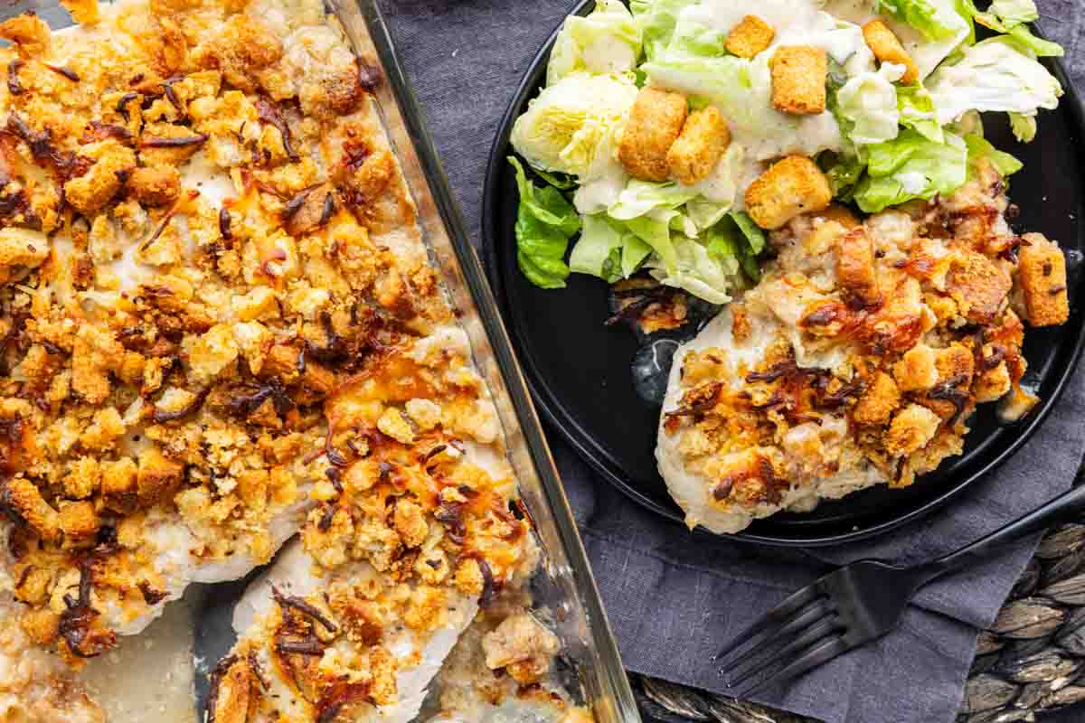 Baked chicken breasts topped with stuffing and cheese in a casserole dish make this easy chicken casserole a comfort food favorite, served on a plate with Caesar salad, croutons, and a touch of Caesar dressing for the perfect Chicken Caesar Bake experience.