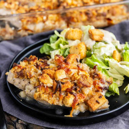 A plate of baked Caesar chicken topped with savory stuffing and melted cheese, served with a side of lettuce salad and crunchy croutons.