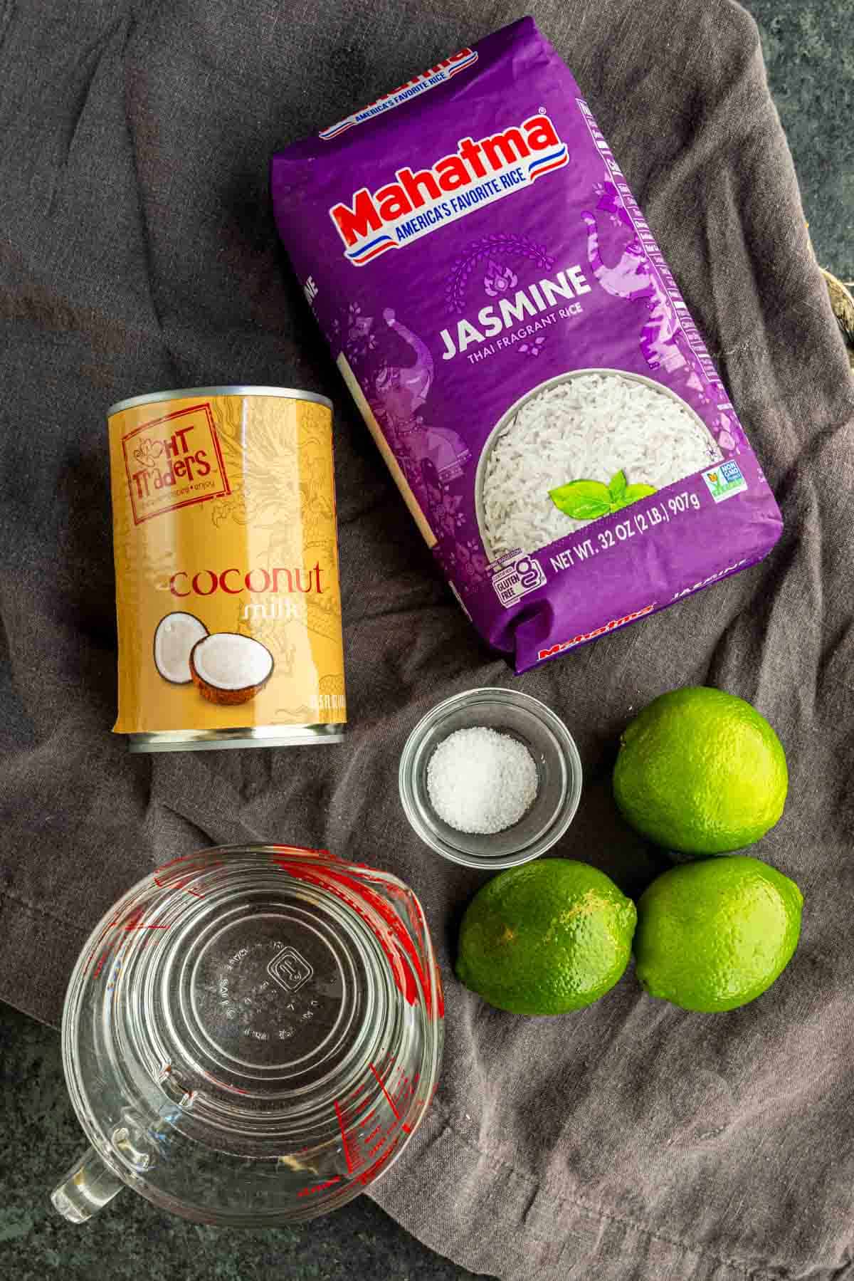 A can of coconut milk, jasmine rice, three limes, a bowl of salt, and a measuring cup of water sit on a dark countertop—perfect for making easy coconut lime rice.