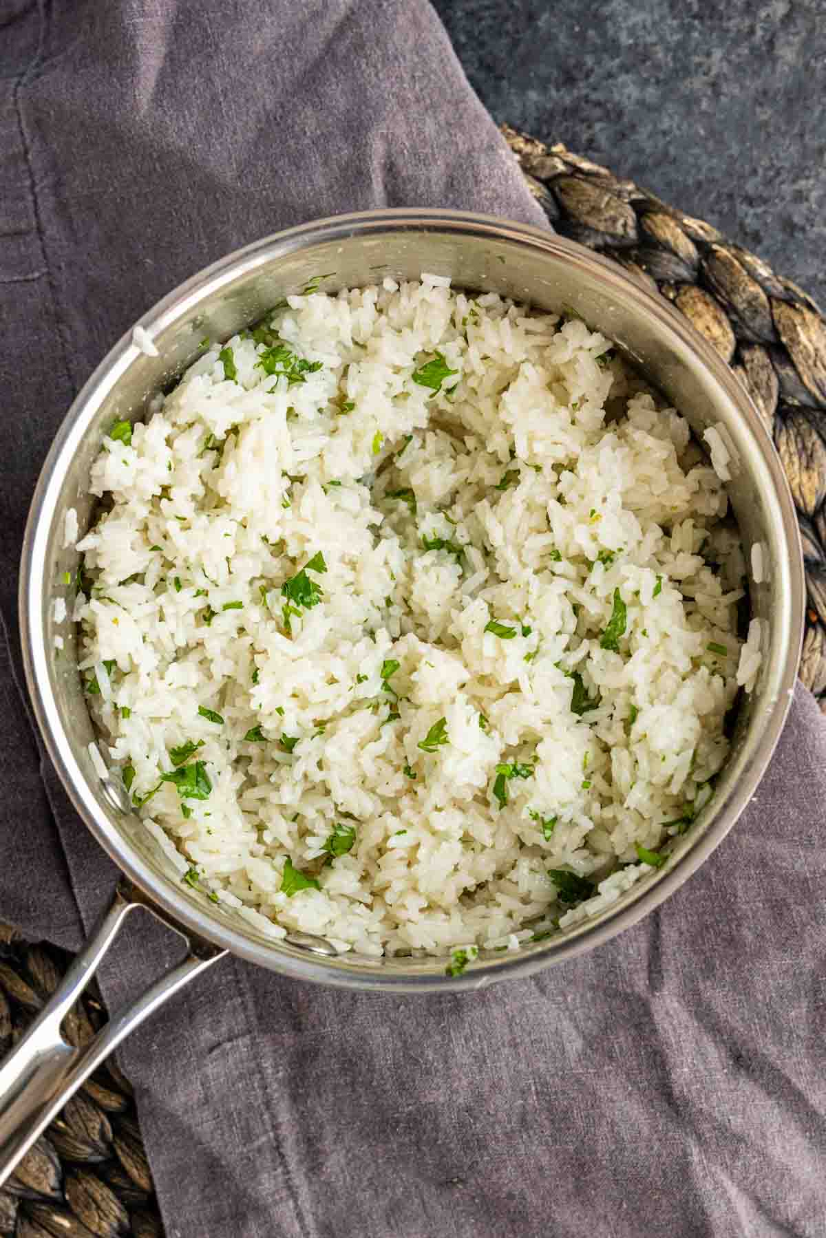 A stainless steel pot filled with easy coconut lime rice, cooked white rice mixed with chopped herbs, placed on a gray cloth and wicker mat.
