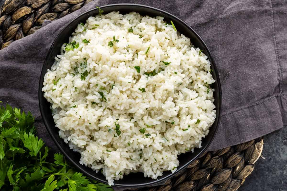 A bowl of easy coconut lime rice garnished with chopped parsley, placed on a dark cloth and woven mat.