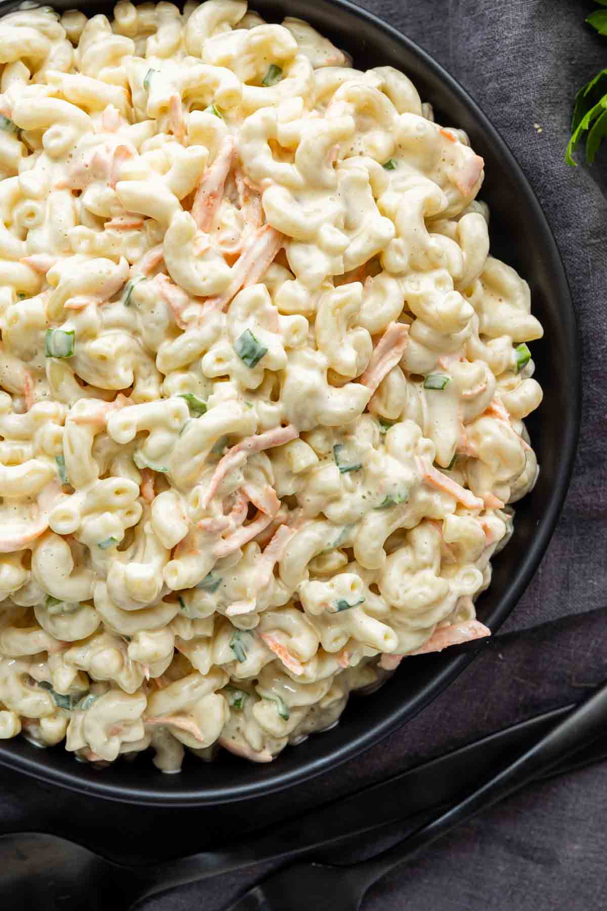 A black bowl filled with creamy Hawaiian macaroni salad—elbow pasta, shredded carrots, and chopped green onions—set on a dark surface. This easy potluck side dish is perfect for any gathering and brings the taste of a classic Hawaiian plate lunch to your table.