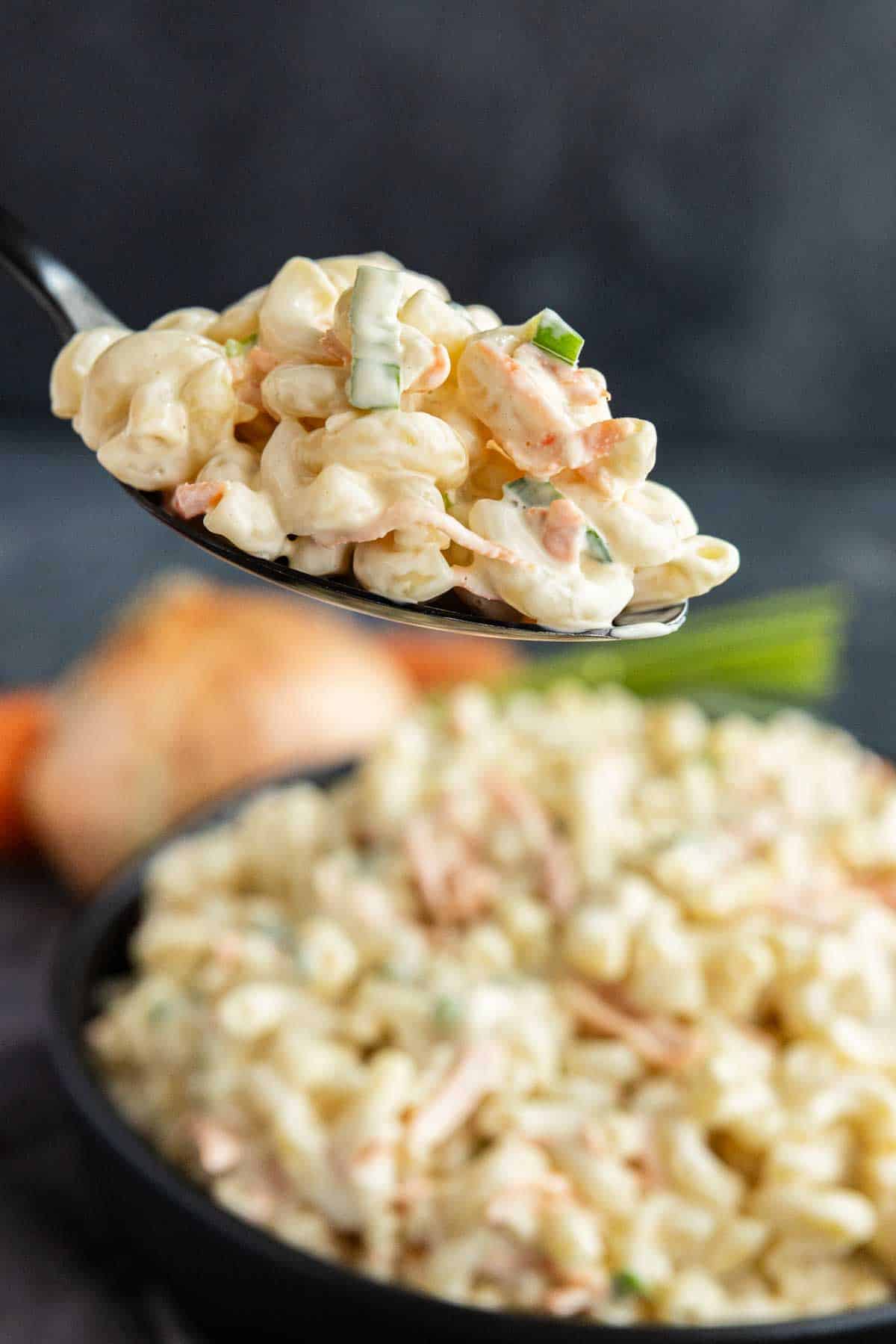 A spoonful of creamy Hawaiian macaroni salad with vegetables is held above a bowl filled with more macaroni salad—an easy potluck side dish that's perfect for any Hawaiian plate lunch.