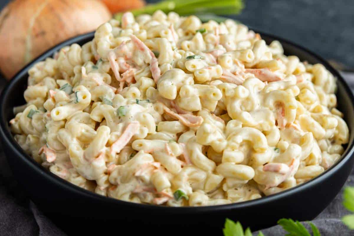 A black bowl filled with creamy Hawaiian macaroni salad featuring shredded carrots and chopped herbs, placed on a dark surface with an onion and greens in the background—an easy potluck side dish that’s sure to please any crowd.