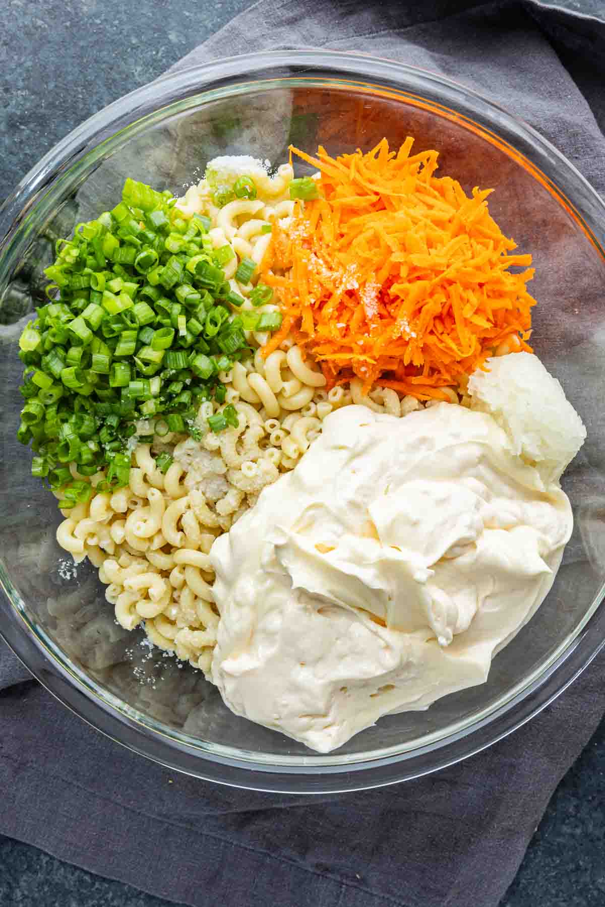 A glass bowl with cooked macaroni, chopped green onions, shredded carrot, mayonnaise, and a scoop of sour cream sits on a dark cloth—a classic Hawaiian macaroni salad perfect as an easy potluck side dish or for serving alongside your favorite Hawaiian plate lunch.