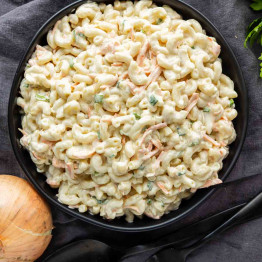 A black bowl filled with Hawaiian macaroni salad—elbow pasta, shredded carrots, green onions, and creamy dressing—sits on a dark cloth with a yellow onion and utensils nearby, making it an easy potluck side dish perfect for any Hawaiian plate lunch.
