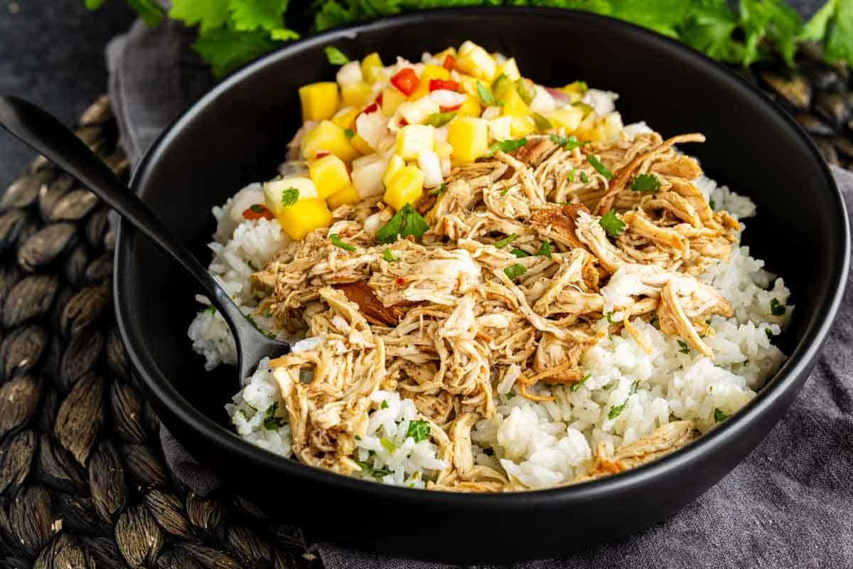 A black bowl filled with shredded chicken over white rice, topped with diced mango salsa, with a black spoon on the side.