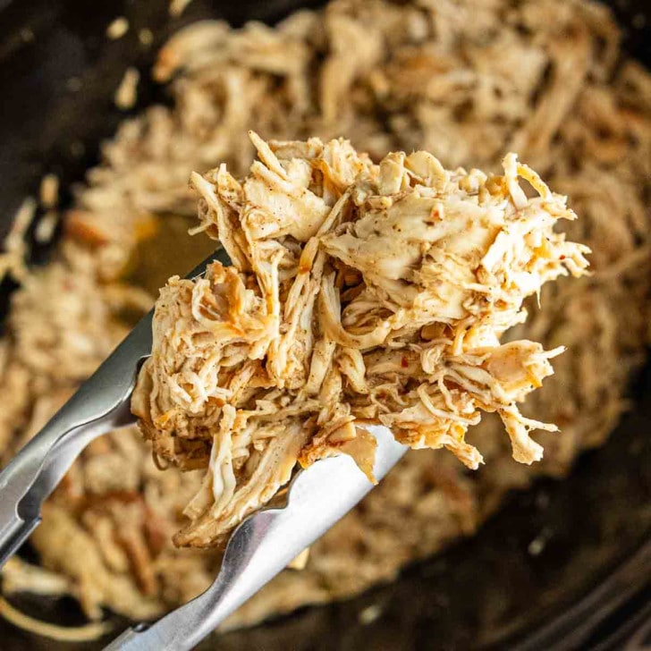 Metal tongs hold shredded cooked chicken above a slow cooker filled with more shredded chicken.