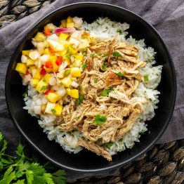 A black bowl with white rice, tender slow cooker jerk chicken, and a colorful fruit salsa on top, placed on a dark fabric surface.
