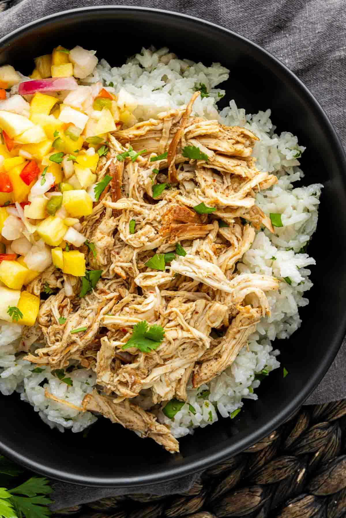 A black bowl filled with white rice, shredded chicken, and a colorful diced fruit salsa, garnished with fresh herbs.