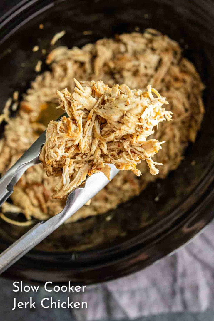 Tongs hold shredded jerk chicken above a slow cooker filled with more shredded chicken. Text at bottom reads "Slow Cooker Jerk Chicken.