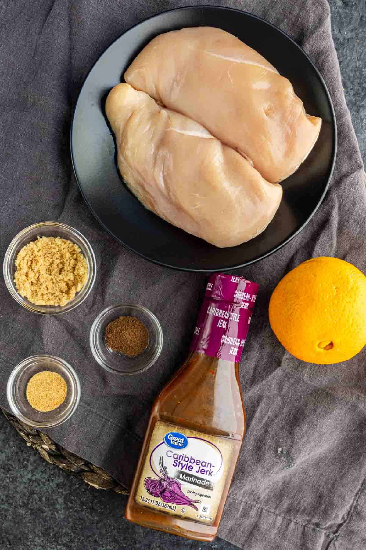 Two raw chicken breasts on a black plate, a bottle of Caribbean-style jerk marinade, an orange, brown sugar, allspice, and garlic powder arranged on a gray cloth.