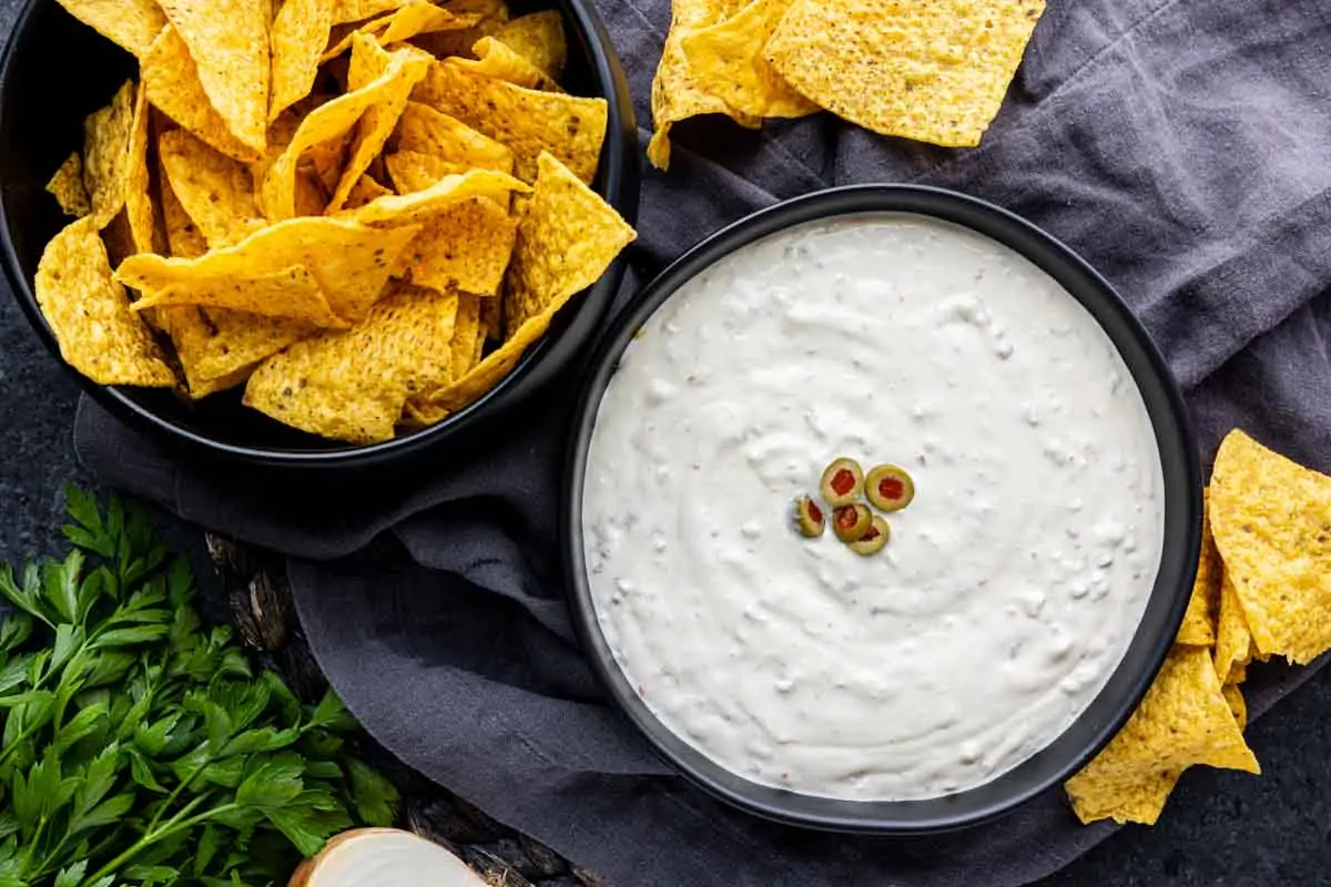 A bowl of tortilla chips next to a bowl of creamy Hamlin's White dip garnished with sliced green olives, placed on a dark cloth with parsley nearby.