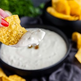 A hand holding a tortilla chip dipped in creamy white sauce, with a bowl of dip and more chips in the background.