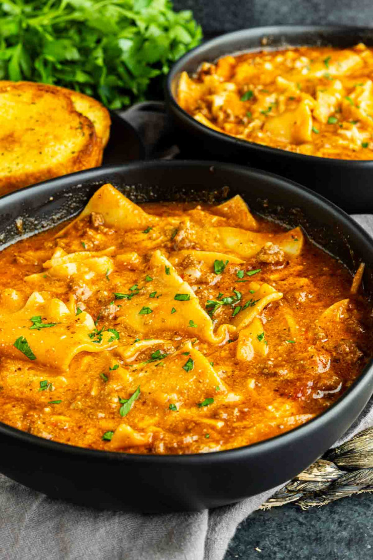 Two black bowls filled with creamy lasagna soup topped with herbs, with a slice of toasted bread and fresh parsley in the background.