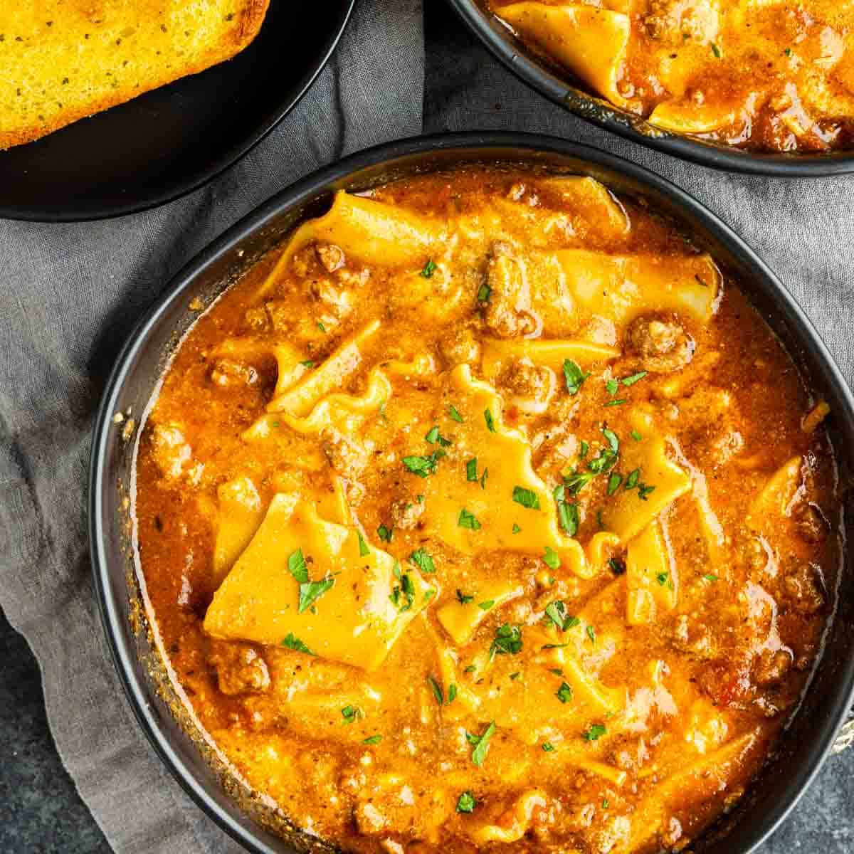 Bowl of lasagna soup with broken pasta pieces, ground meat, tomato sauce, and herbs, served on a dark surface with a piece of bread on the side.