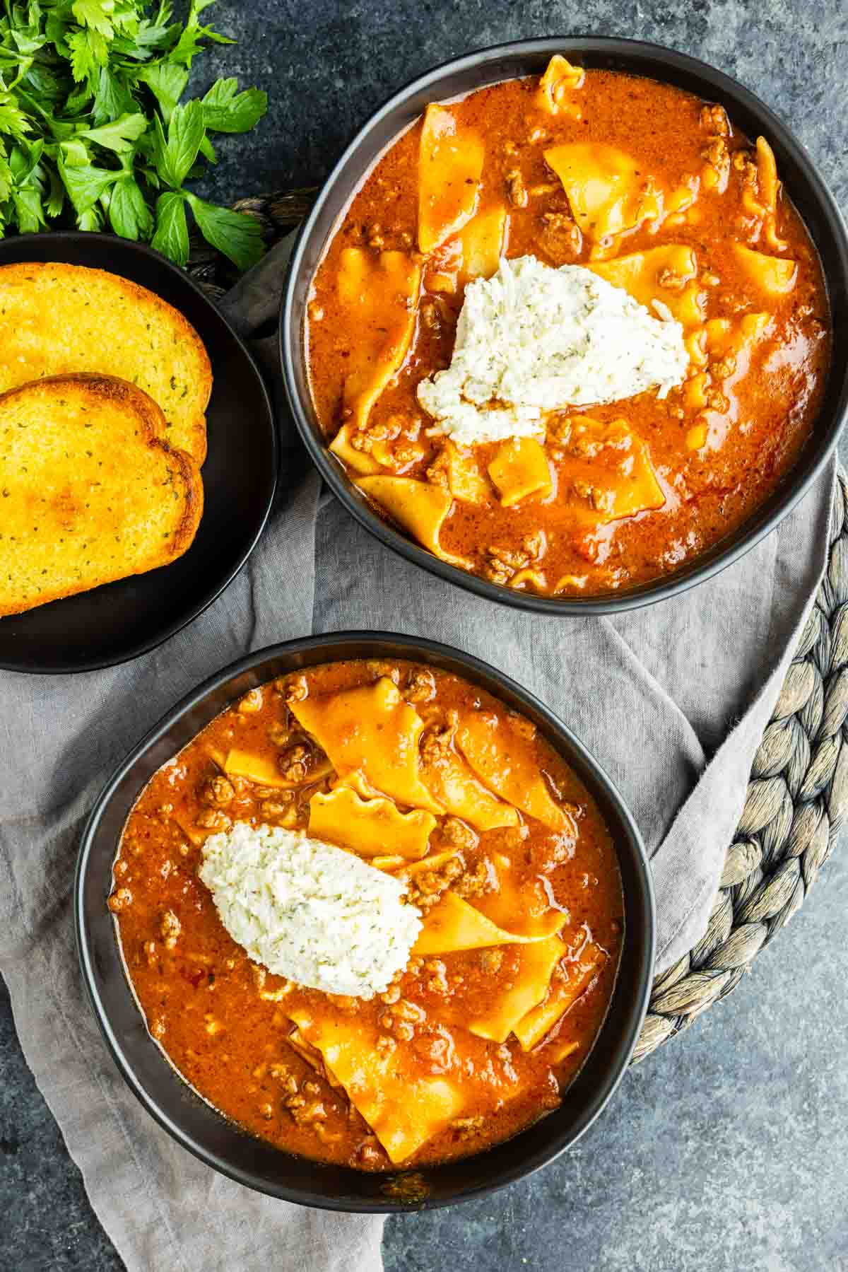 Two bowls of lasagna soup with ricotta cheese on top, served next to slices of toasted bread and a bunch of fresh parsley.