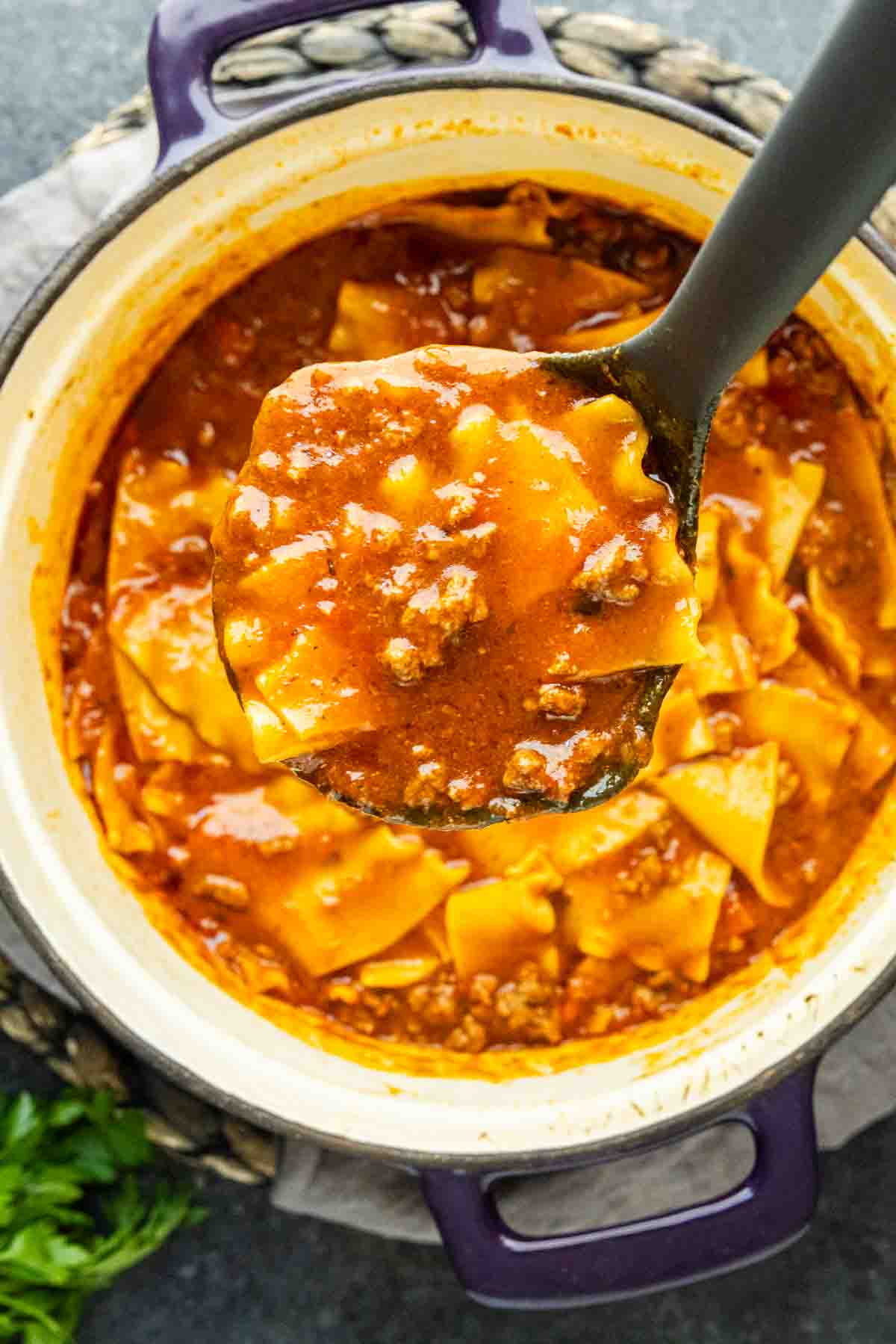 A ladle lifts a serving of lasagna soup with ground meat and pasta from a pot filled with the tomato-based soup.
