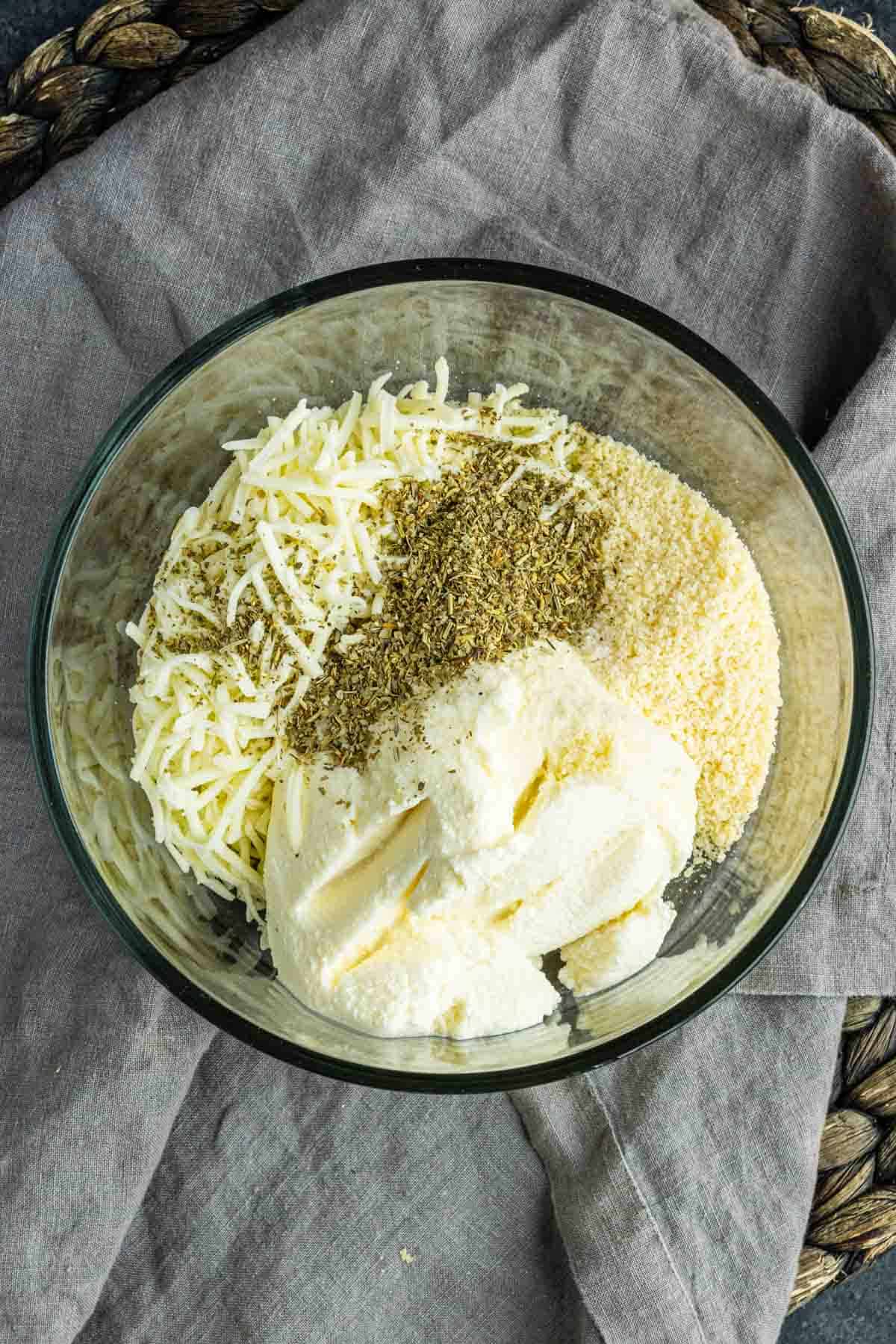 A glass bowl with ricotta cheese, shredded mozzarella, grated parmesan, and Italian seasoning on a gray cloth.