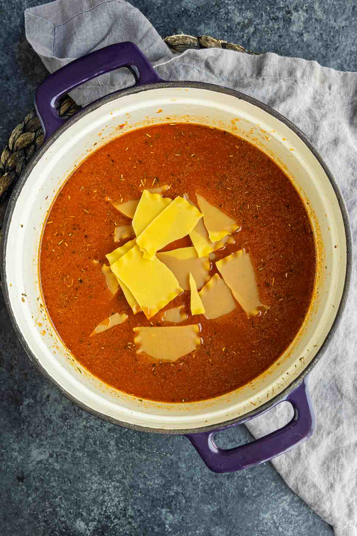 A pot of tomato-based soup with broken lasagna noodles and shredded cheese on top, sitting on a gray cloth on a dark surface.
