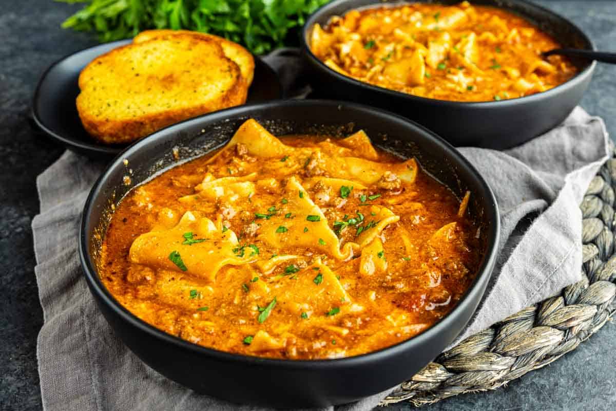 Two black bowls of lasagna soup topped with herbs are set on a gray napkin, with a plate of toasted bread in the background.