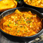 A black bowl of one pot lasagna soup with ground meat, lasagna noodles, tomato sauce, and herbs, with a slice of toasted bread in the background.