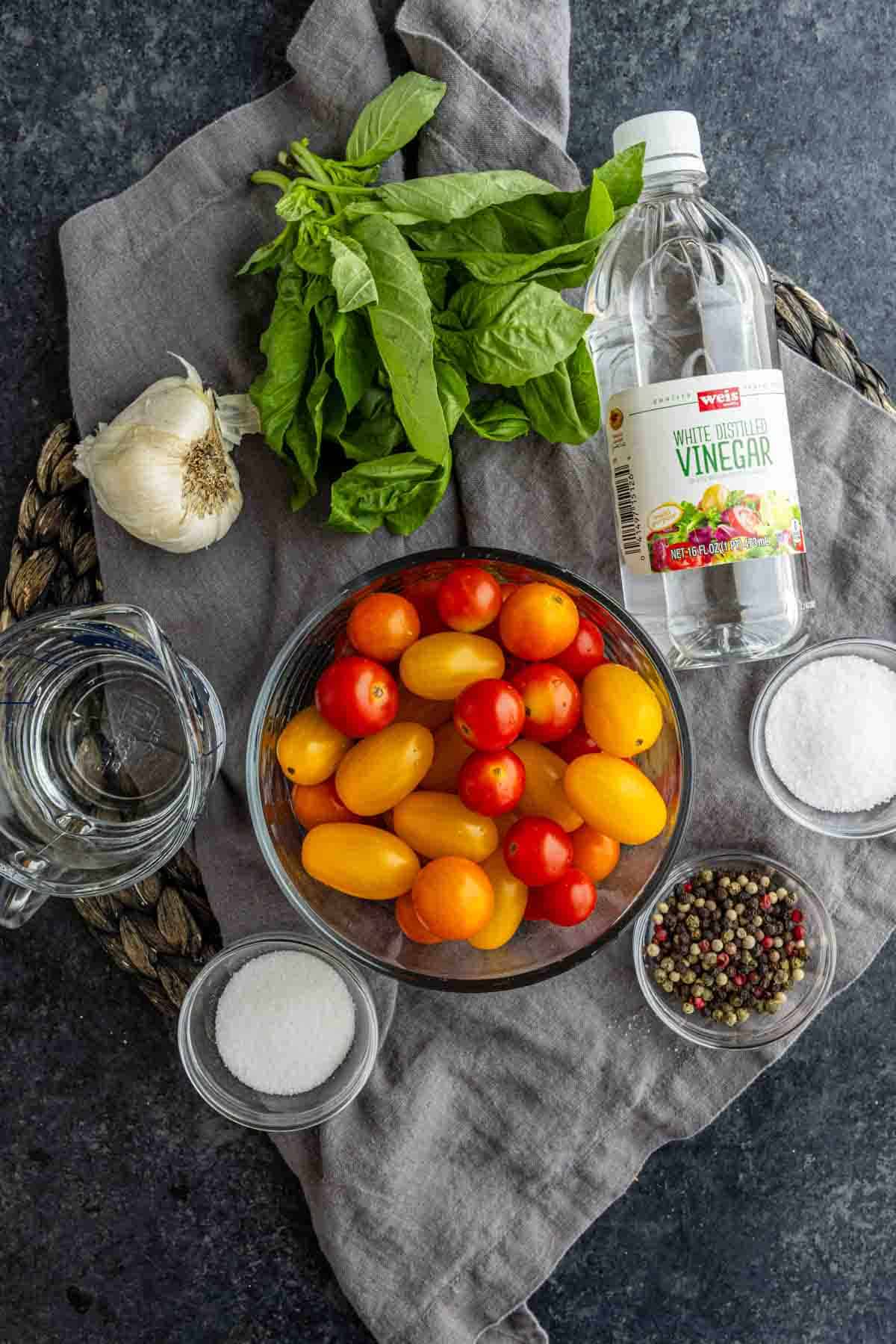A bowl of cherry tomatoes, basil, garlic, white vinegar, water, salt, sugar, and peppercorns are arranged on a dark surface with a gray cloth—everything you need for a quick pickle recipe.