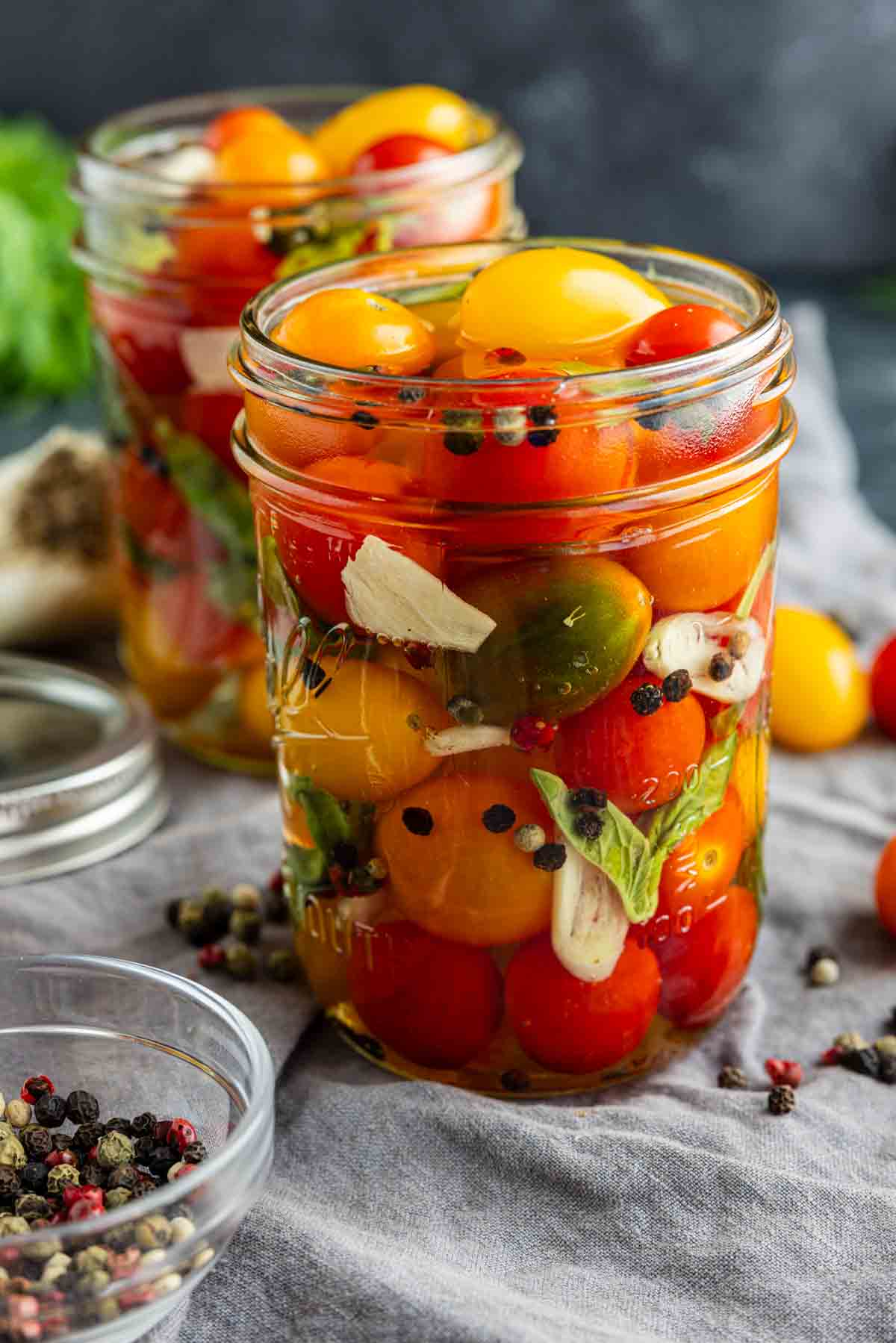 A glass jar filled with colorful pickled tomatoes, herbs, garlic slices, and peppercorns in brine illustrates a quick pickle recipe in action, with another jar and loose ingredients enticingly arranged in the background.