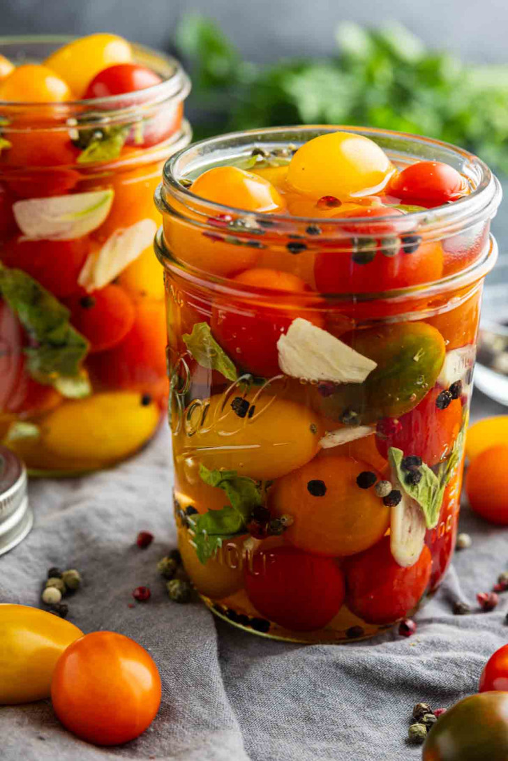 A glass jar filled with pickled cherry tomatoes, garlic slices, peppercorns, and herbs, sitting on a gray cloth with loose tomatoes nearby.