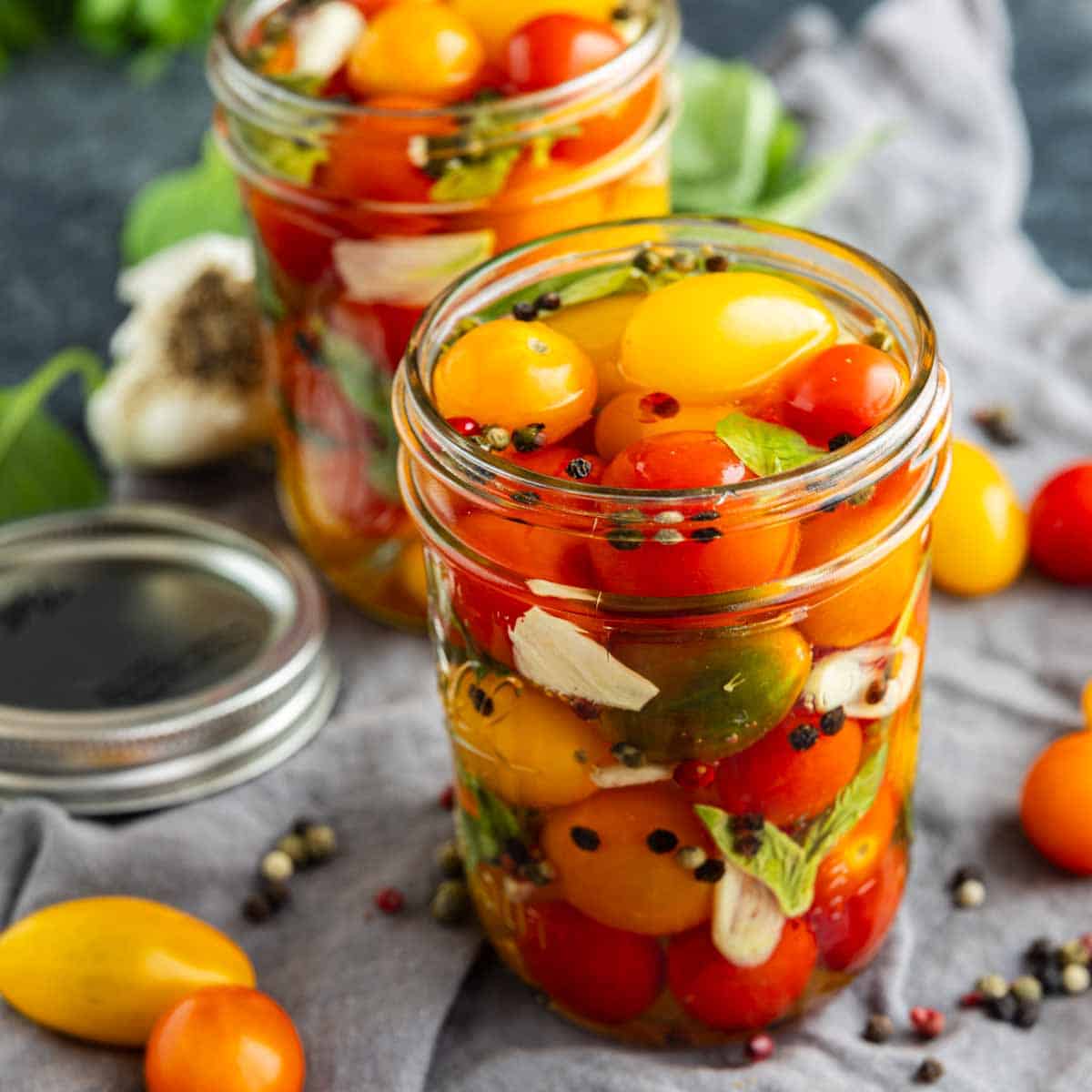Two glass jars filled with pickled cherry tomatoes, herbs, garlic, and peppercorns sit on a gray cloth with scattered tomatoes and spices around them—a perfect snapshot of a quick pickle recipe that showcases how to pickle cherry tomatoes at home.