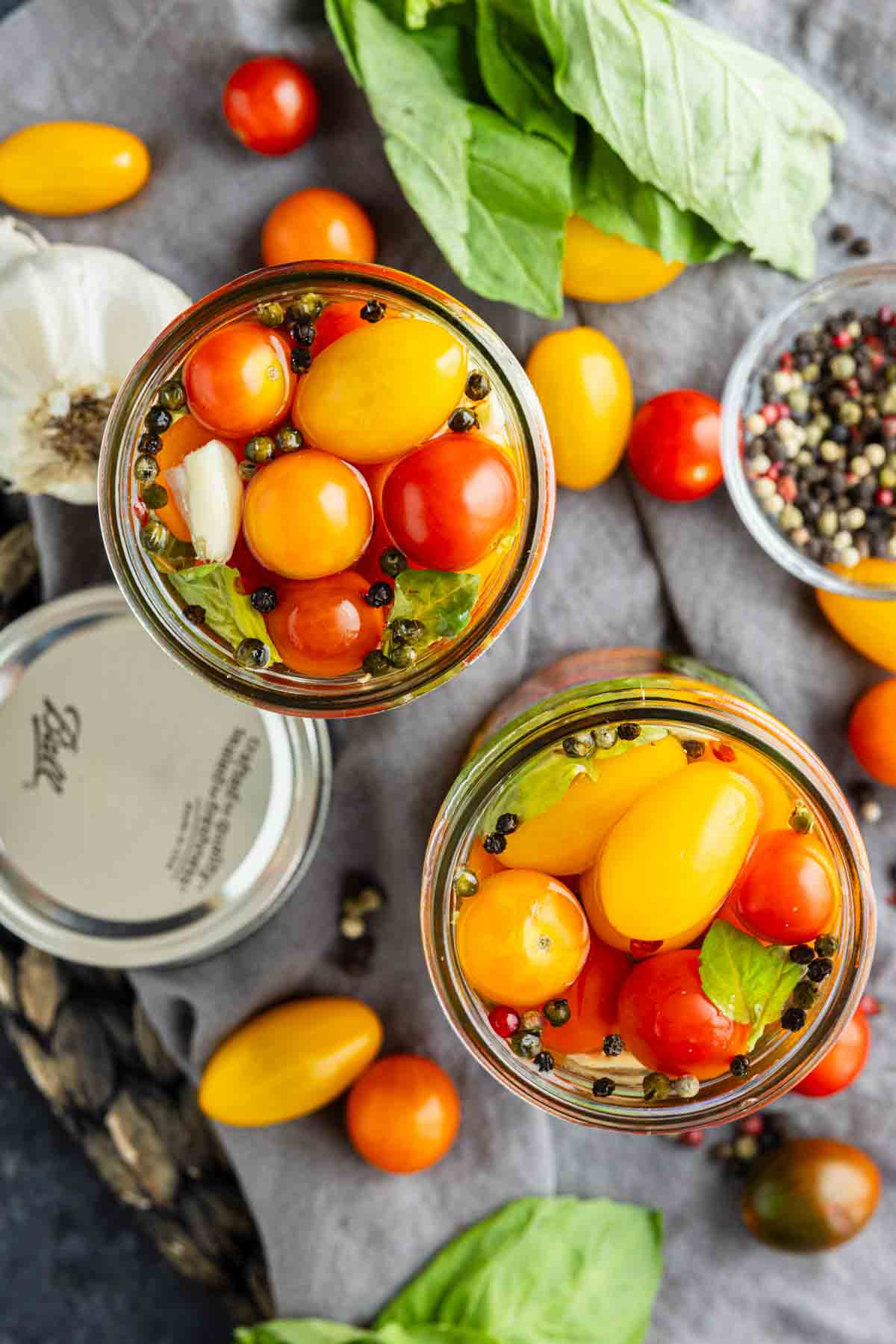 Two glass jars filled with vibrant red and yellow pickled cherry tomatoes, basil leaves, garlic, and peppercorns, surrounded by fresh tomatoes, basil, pepper, and a jar lid on a gray cloth—a perfect inspiration if you’re curious about how to pickle cherry tomatoes at home.