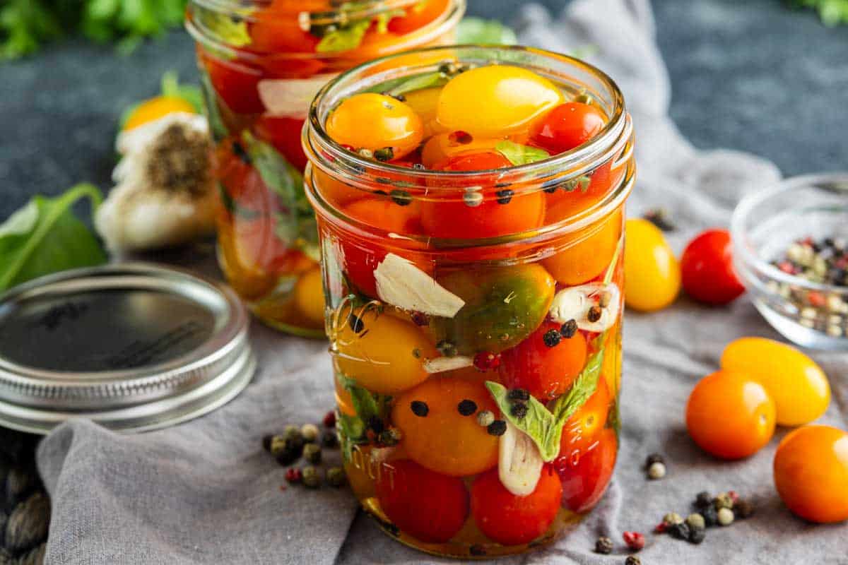Two glass jars filled with pickled cherry tomatoes, herbs, garlic slices, and peppercorns, with loose tomatoes and peppercorns scattered nearby. One jar lid is off and resting on the table—showcasing exactly how to pickle cherry tomatoes at home.