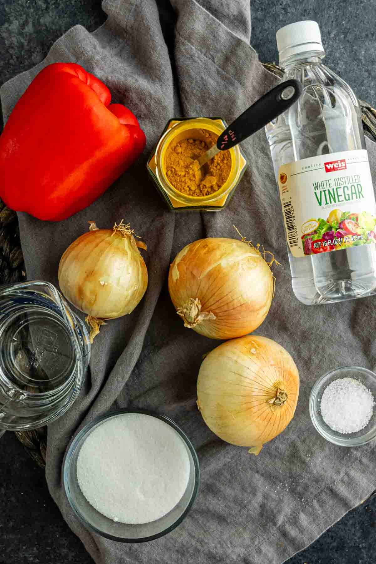 A red bell pepper, three yellow onions, curry powder, white vinegar, salt, sugar, and a measuring cup of water are arranged on a gray cloth.