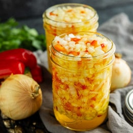 Two glass jars filled with chopped vegetables in yellow brine, with onions, red pepper, and herbs in the background on a gray cloth.
