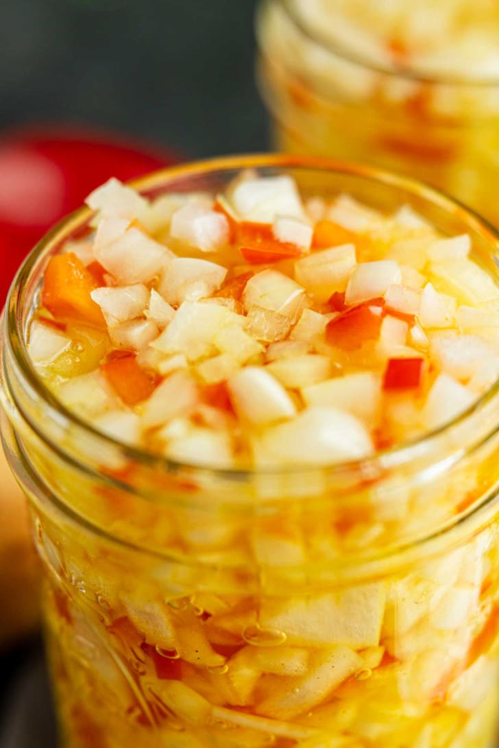 A close-up view of a glass jar filled with chopped onions and red peppers in a yellow liquid, likely a pickling brine.